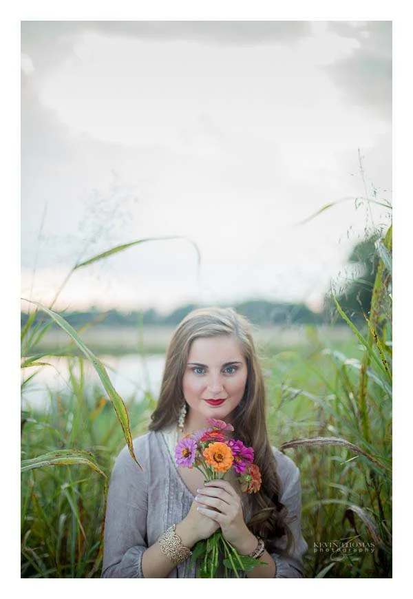 Young woman with long brown hair and red lipstick holding a bouquet of colorful flowers, standing outdoors among tall green grasses with a body of water and cloudy sky in the background.