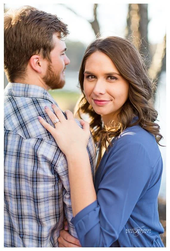 A young couple standing outdoors, facing each other closely, with the woman looking at the camera and smiling. The man is facing sideways, with his eyes closed, and has his arm around her. The woman has long brown hair and wears a blue blouse, while 