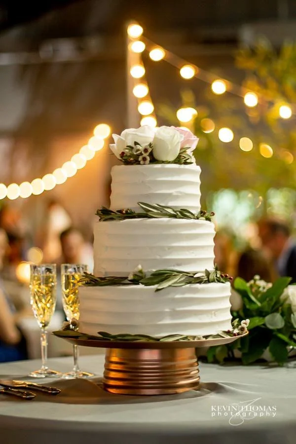 A three-tier white wedding cake decorated with white roses and greenery, set on a table with gold utensils and champagne glasses, with blurred string lights and guests in the background.