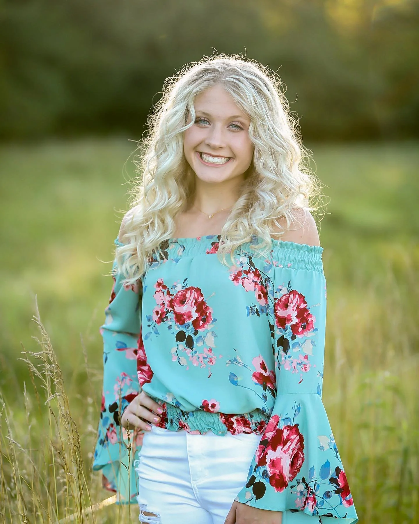 A young woman with long, curly blonde hair and blue eyes, smiling outdoors in a grassy field, wearing a turquoise off-the-shoulder blouse with pink and red floral patterns and white ripped jeans.