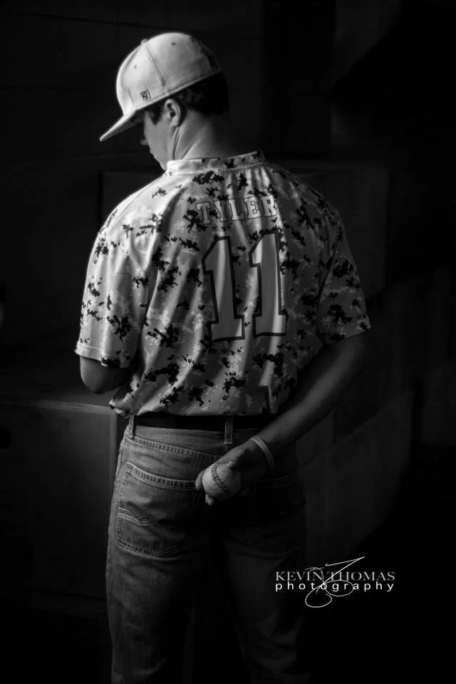 A person seen from behind wearing a baseball cap, a sports jersey with the name 'Tiller' and the number 11, and jeans. The person is standing indoors, holding a baseball in their left hand behind their back.