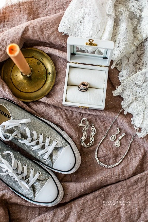 A jewelry box with rings, a pair of earrings, and a necklace; a pair of sequined sneakers; lace fabric; and a candle holder with a candle on a soft, pinkish fabric surface.