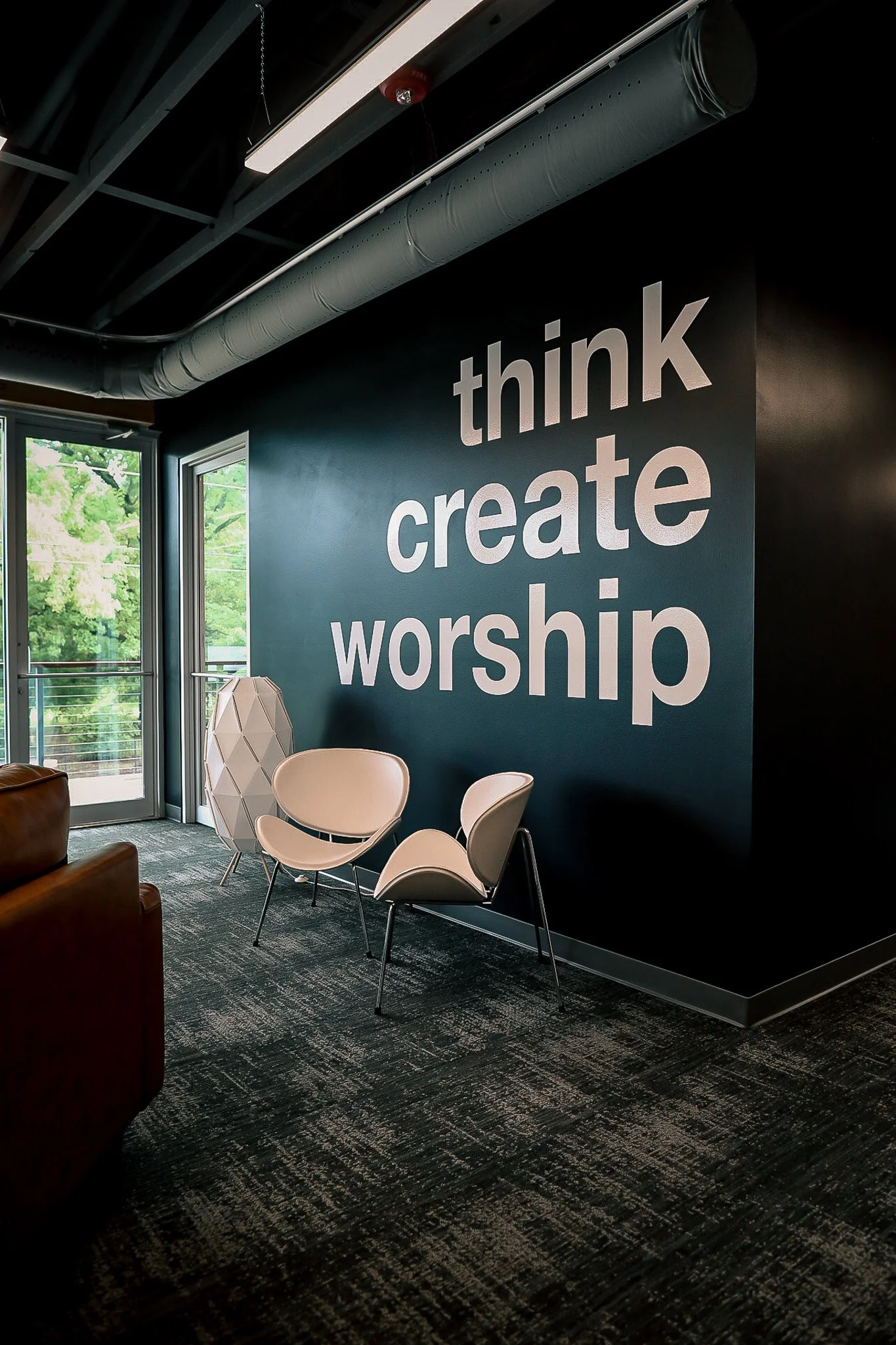Interior of a room with a dark blue wall displaying the words "think create worship" in large white letters. There are three beige chairs, a decorative white lamp, and a door leading outside with trees visible through the glass.