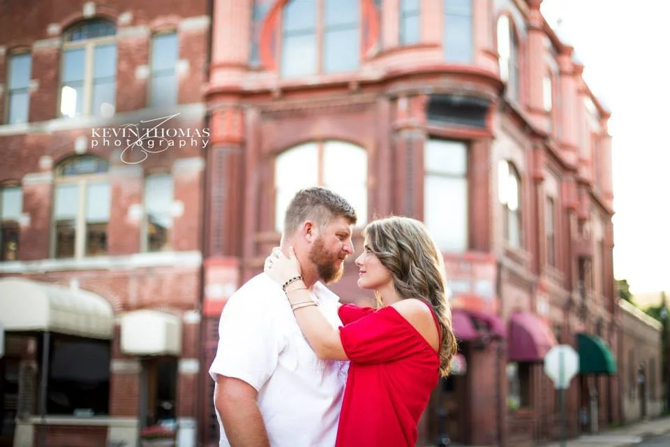 A couple stands close together in front of a historic brick building, looking into each other's eyes, with the woman holding the man's face.