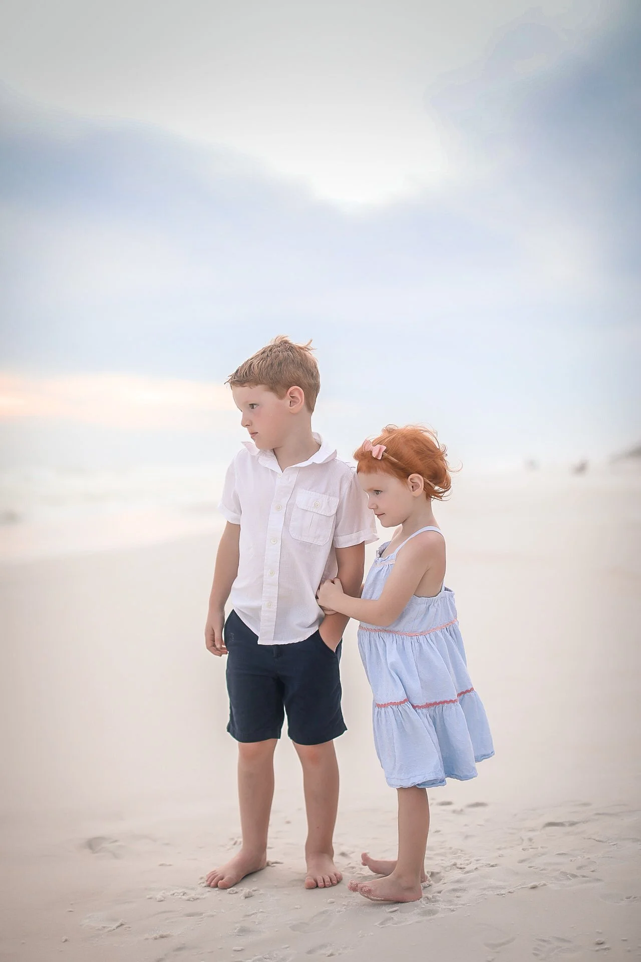A boy and girl standing on the beach with a cloudy sky in the background. The girl is holding the boy's arm, and they are barefoot on the sand.