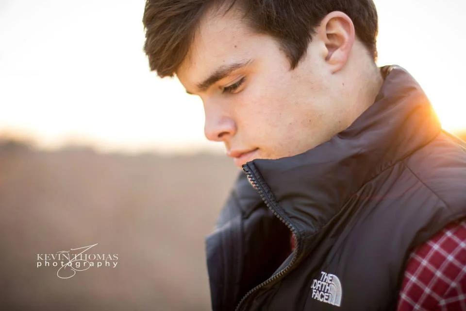 Close-up of a young man with brown hair, wearing a black North Face jacket and red plaid shirt, looking down outdoors at sunset.