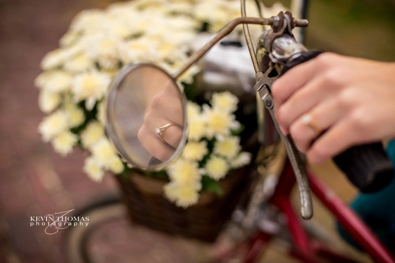 Close-up of a person's hand holding handlebars of a bicycle with a small mirror reflecting a ring on their finger, with a basket of white flowers in the background.