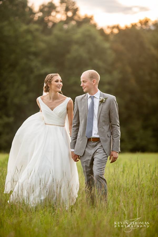 A bride and groom walking hand in hand through a grassy field at sunset, smiling at each other. The bride wears a white wedding gown, and the groom is in a gray suit with a blue tie and boutonniere.
