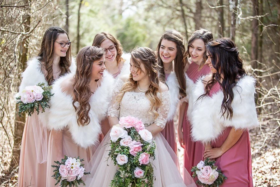 A bride and six bridesmaids smiling and gathering in a forest, with the bridesmaids wearing pink dresses and white faux fur stoles, holding bouquets of pink and white flowers.