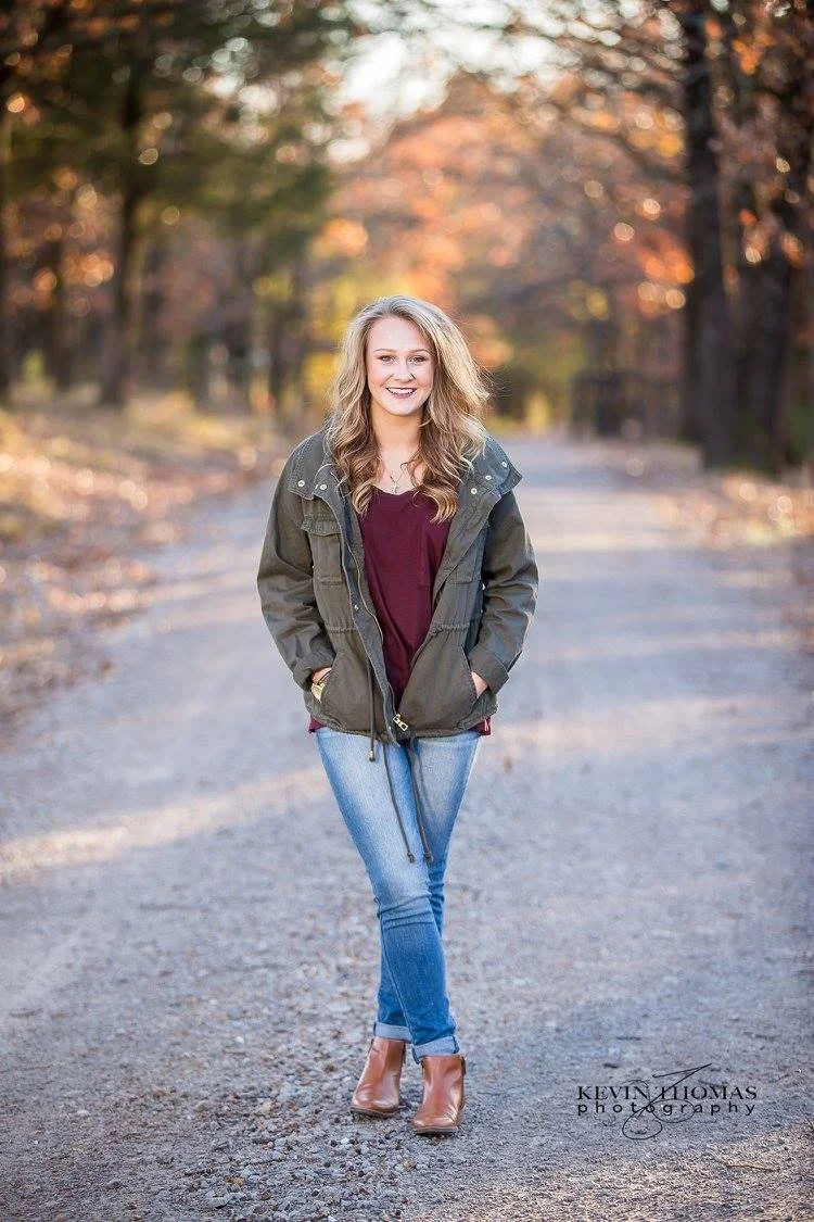 A young woman smiling and walking on a tree-lined dirt road during autumn, wearing a green jacket, maroon top, blue jeans, and brown ankle boots.