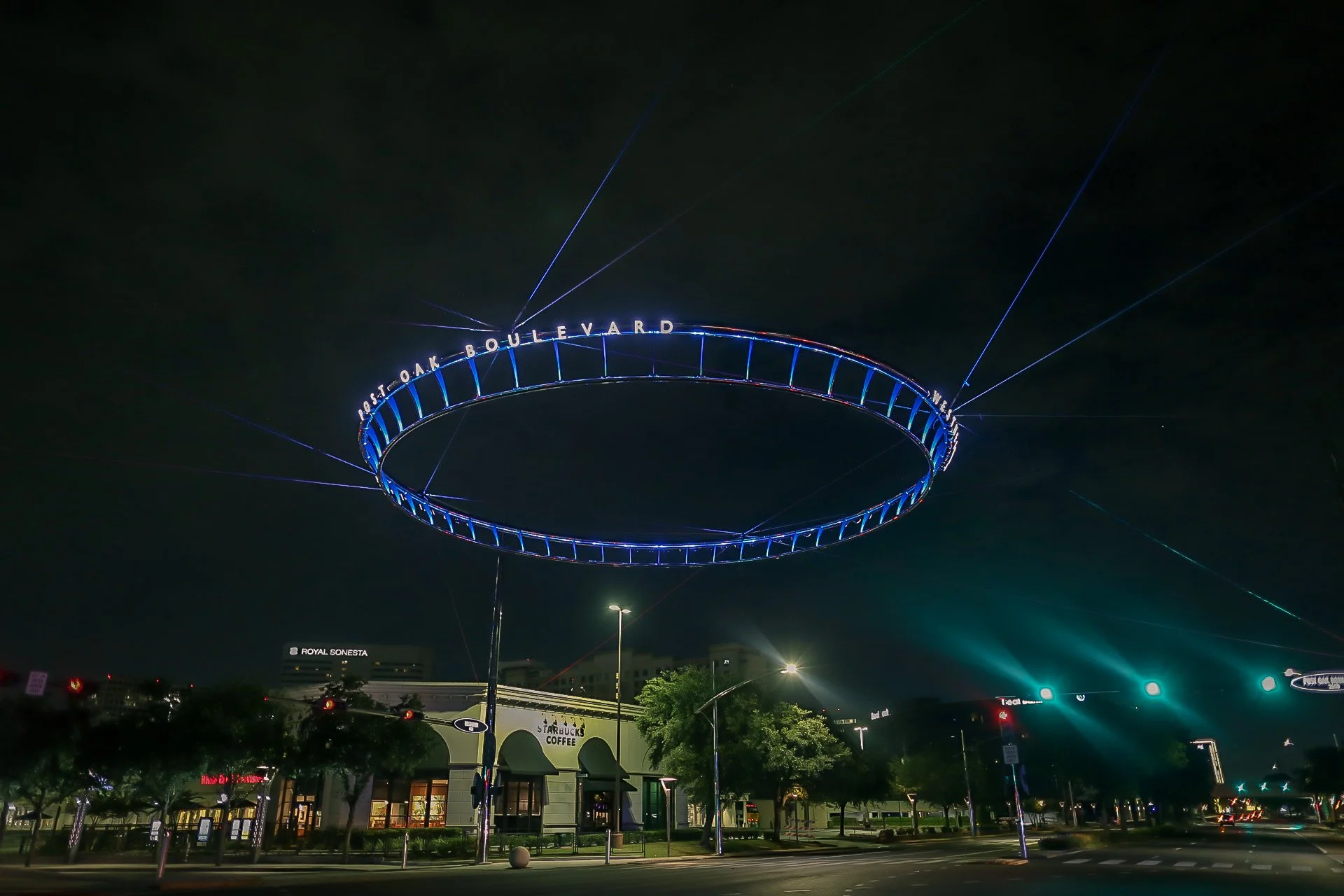 Night view of a large illuminated circular ring structure with blue lights and the words 'East Oak Boulevard West' on it, floating above a city street with trees, streetlights, and buildings including Starbucks Coffee and Royal Sonesta.
