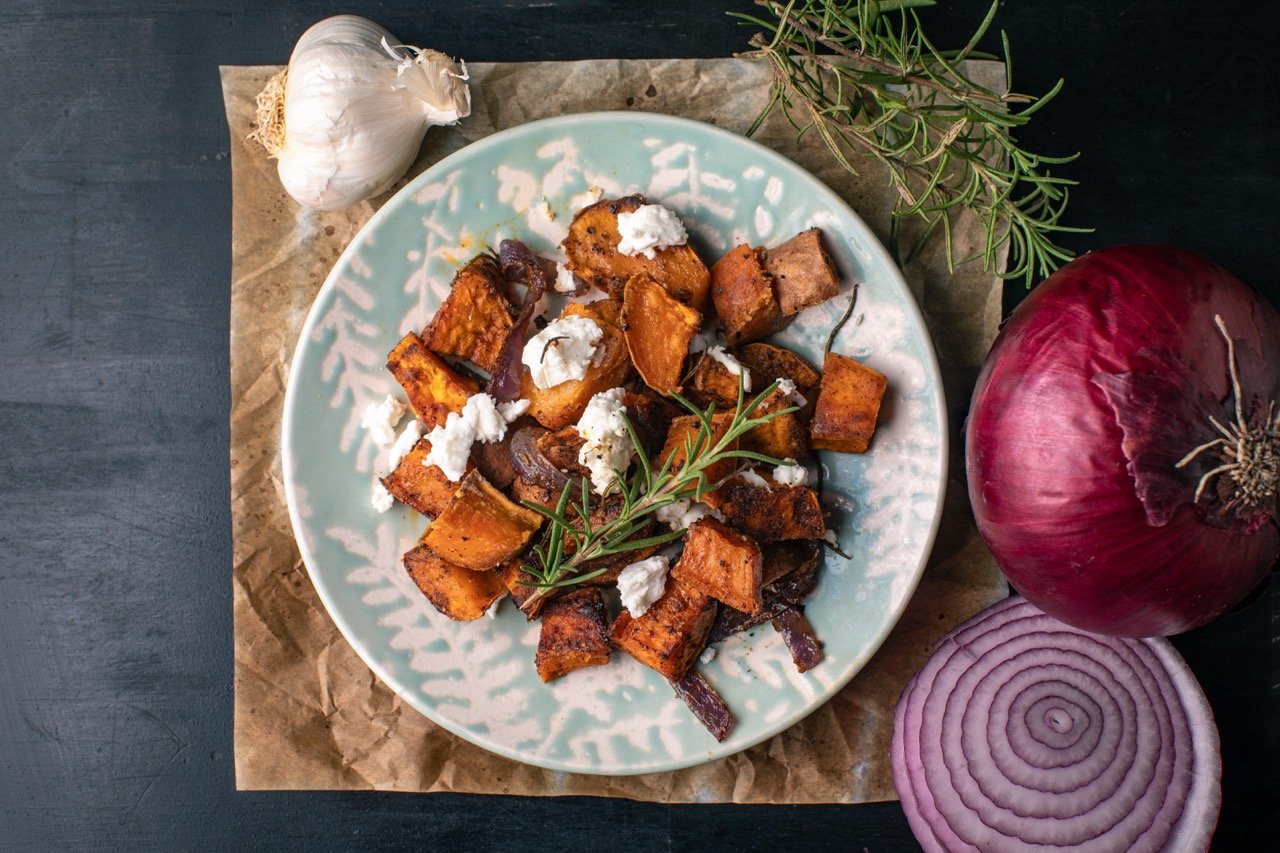 A plate of roasted sweet potatoes topped with crumbled cheese and fresh rosemary, surrounded by a whole garlic bulb, a red onion, and a sliced red onion on a dark surface.