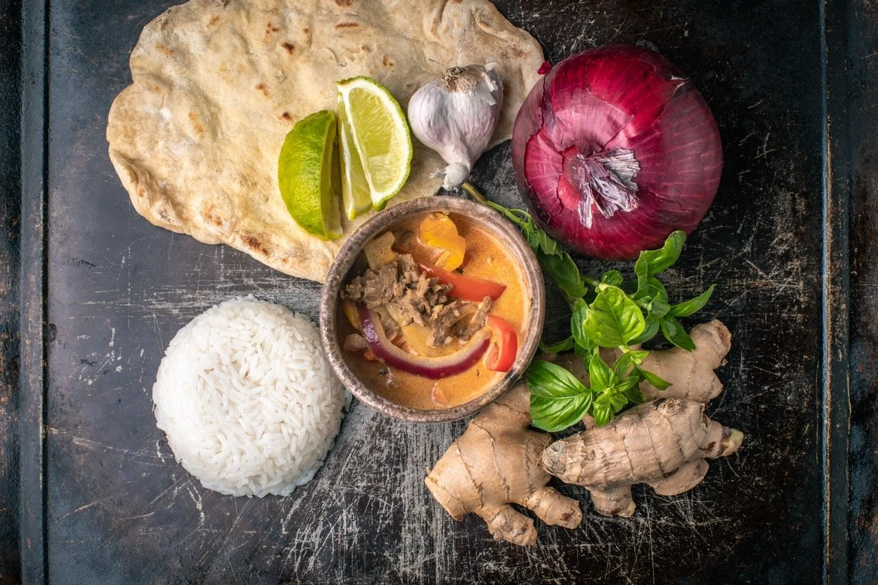 A serving of Thai red curry with rice, lime wedges, garlic, red onion, ginger, fresh basil, and a piece of flatbread on a dark surface.