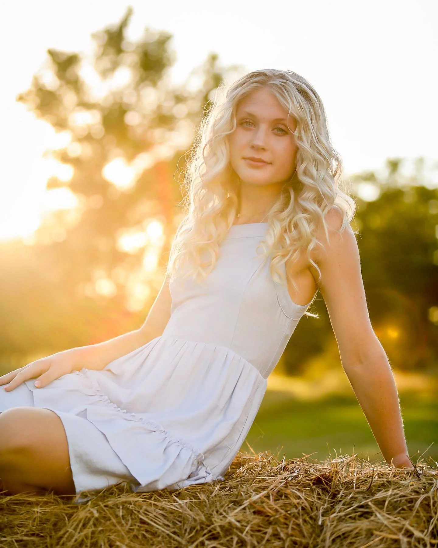 A young woman with blonde curly hair sitting on hay in a white dress outdoors during sunset.
