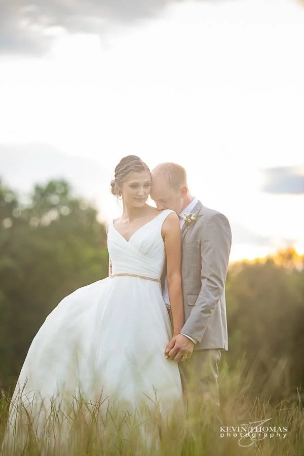 A bride and groom standing together in a field during sunset, with the bride wearing a white wedding dress and the groom in a gray suit.