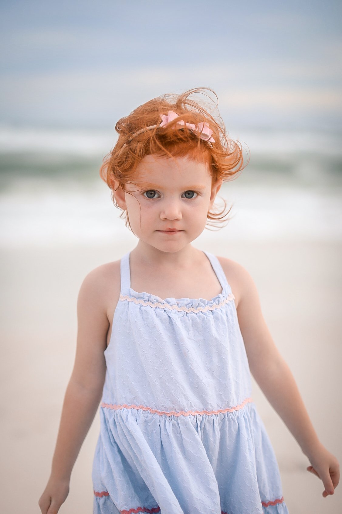 A young girl with red hair and blue eyes standing on a beach with the ocean in the background, wearing a light blue dress.