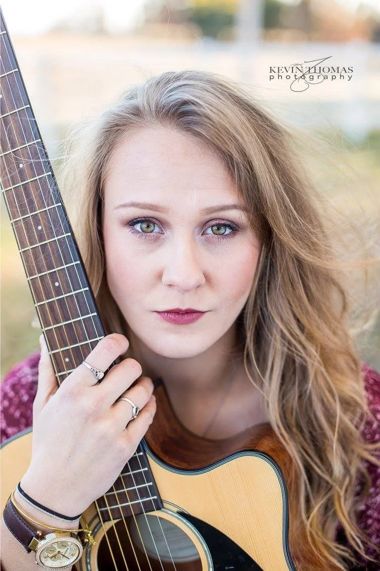 A young woman with wavy blonde hair and blue eyes holding an acoustic guitar close to her face. She is wearing a maroon top and jewelry, including rings and a watch.