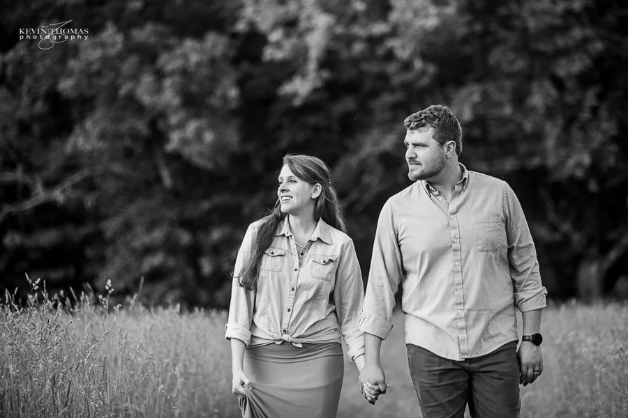A couple holding hands walking in a field with trees in the background in black and white.