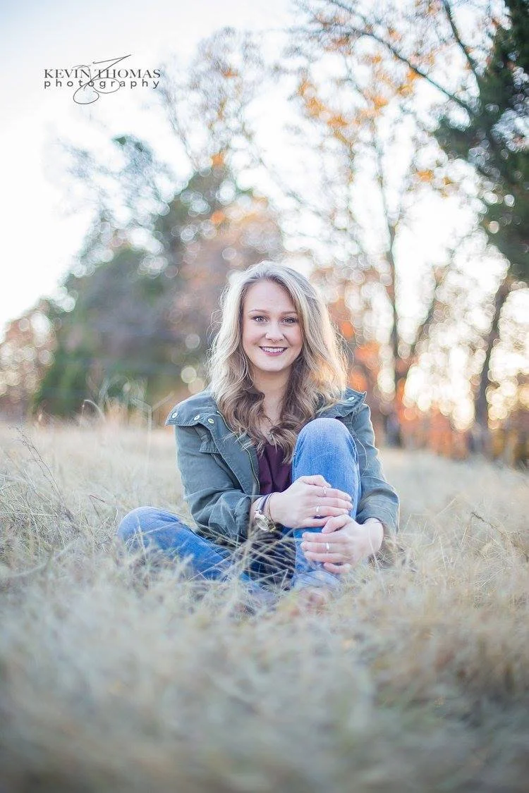Young woman sitting outdoors on dry grass in autumn, smiling at the camera, wearing a green jacket and jeans, with a blurred background of trees with fall foliage.