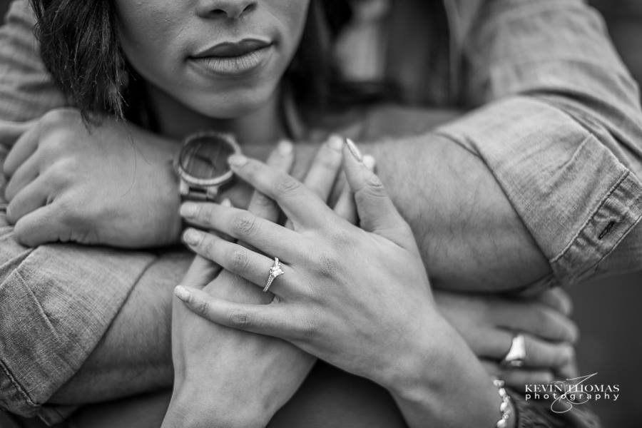 Close-up of a woman with an engagement ring and a watch, being embraced from behind by a person's arms, in black and white.