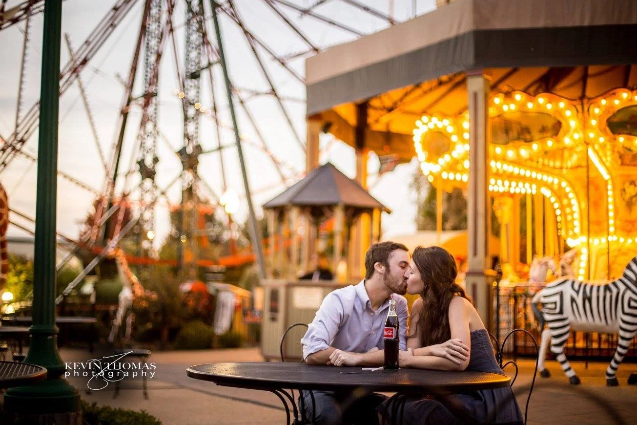 A man and woman are sharing a kiss at an outdoor amusement park table with a soda bottle. In the background, there is a carousel with bright lights and a zebra figure.