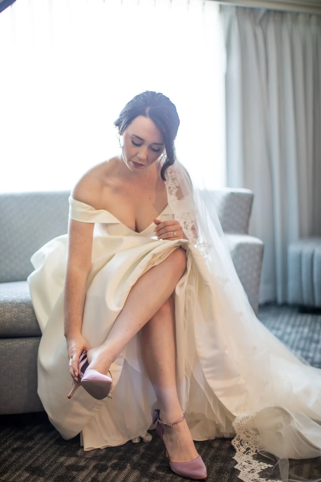 A bride in a white wedding dress putting on pink high-heeled shoes in a hotel room.