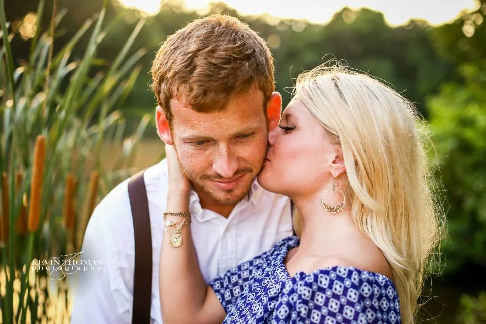 A woman with blonde hair is kissing a man on the cheek, with her hand gently holding his face. The man has short, light brown hair and is smiling with his eyes closed. They are outside near greenery and tall plants during sunset.