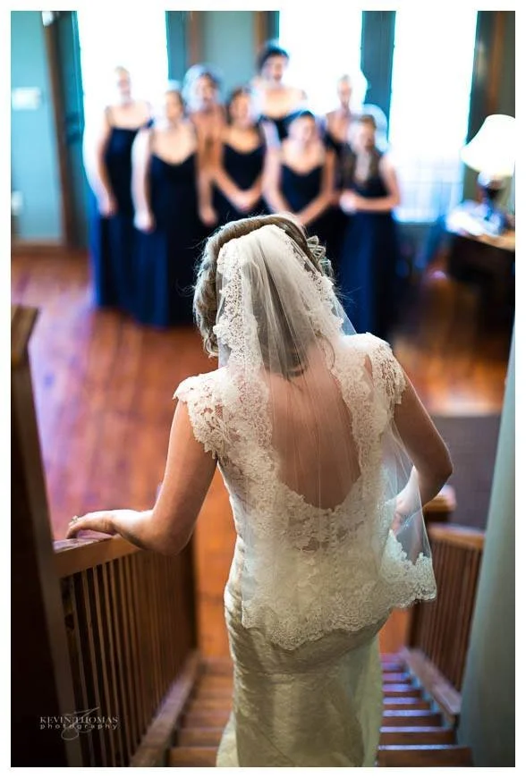 Bride in a lace wedding dress with a veil, ascending stairs and looking towards a group of women in black dresses.