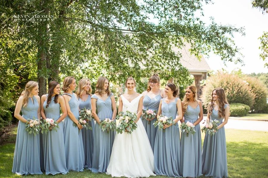 A bride and seven bridesmaids standing outdoors on grass with trees in the background. The bridesmaids are wearing matching light blue dresses, and the bride is in a white gown. They are holding bouquets of flowers and smiling.