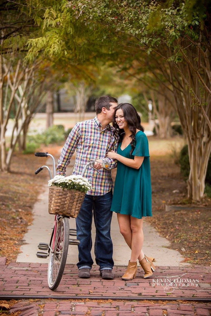 A young couple standing on a brick path in a park, with the man kissing the woman on the cheek. The man holds a bicycle with a basket of white flowers. The woman is smiling, wearing a teal dress and ankle boots. Trees with green foliage form a canopy