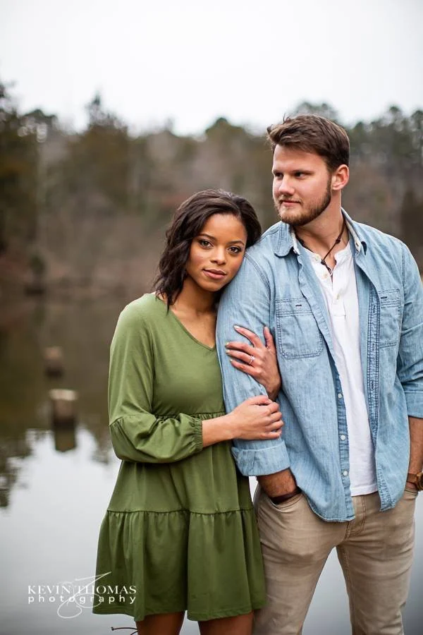 A young woman with dark hair in a loose green dress stands beside a young man with facial hair in a light blue denim shirt and beige pants. They are outdoors near a body of water with trees in the background, looking at each other fondly.