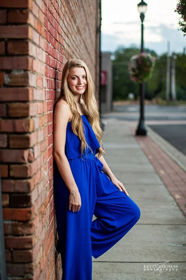 A young woman in a matching blue jumpsuit stands against a brick wall on a city sidewalk, smiling at the camera with her left arm resting on her hip.