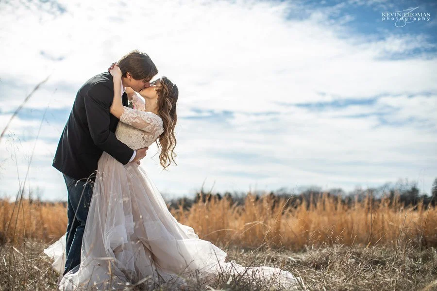 A couple sharing a kiss in a field with tall grass under a partly cloudy sky, the woman in a wedding dress and the man in a black suit.