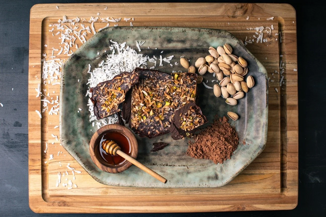 Plate with chocolate bark topped with nuts, shredded coconut, a pile of pistachios, honey with a honey dipper, and cocoa powder on a wooden cutting board.