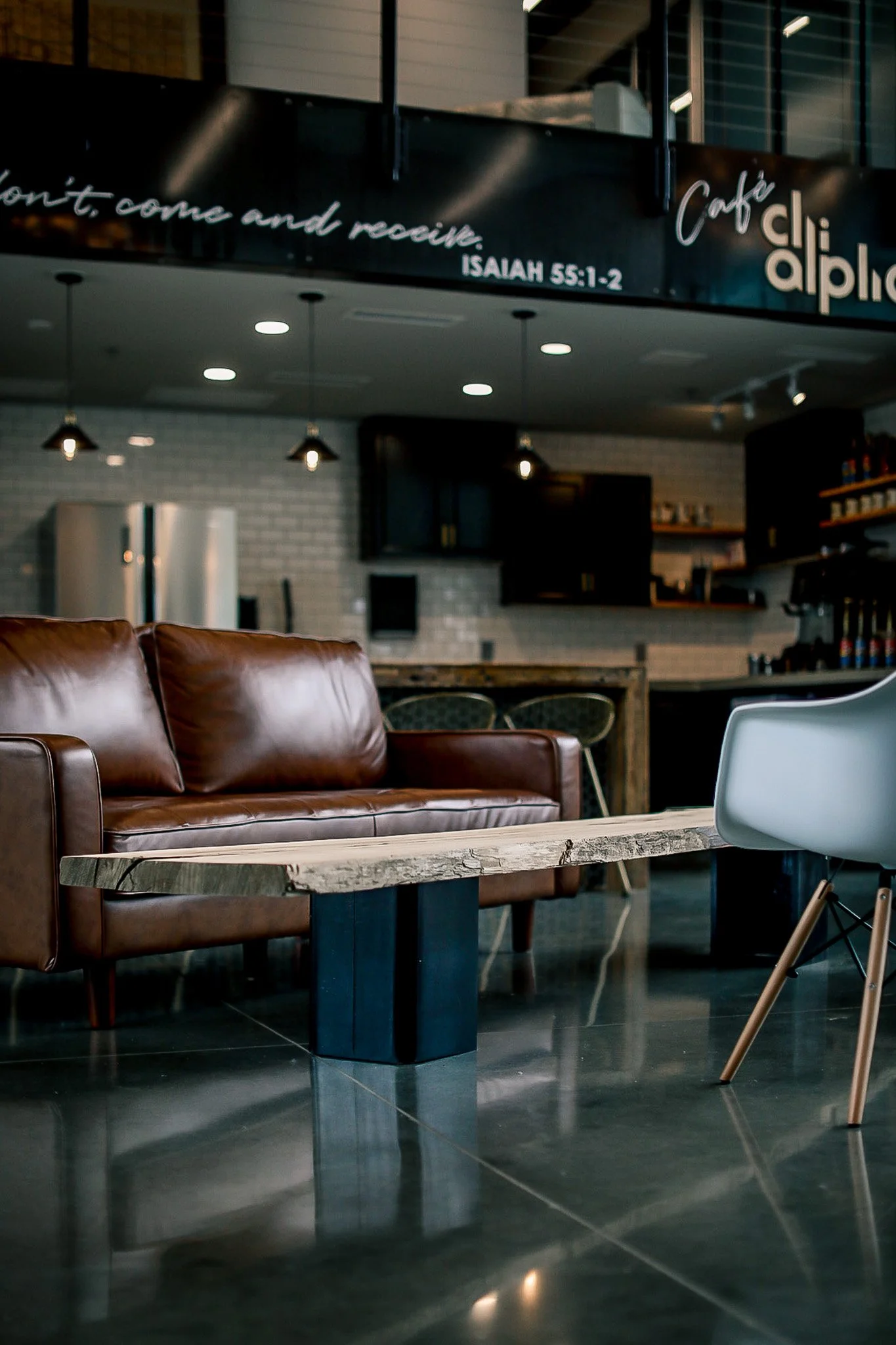 Interior of a modern coffee shop with a brown leather sofa, a rustic wooden coffee table, and a white modern chair. In the background, there is a kitchen area with a refrigerator and shelves, illuminated by overhead lighting.