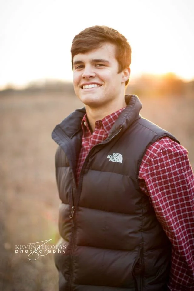 Young man smiling outdoors at sunset, wearing a red checkered shirt and a black North Face puffer vest.