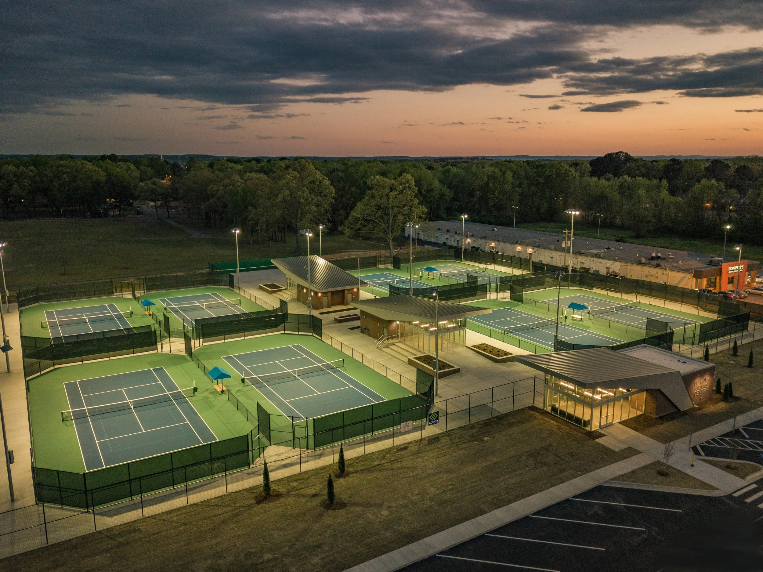 An outdoor tennis court complex with multiple lit courts, some with blue and green canopies, surrounded by a fence, at sunset or dusk. There are parking lots and a building with a lit entrance nearby.
