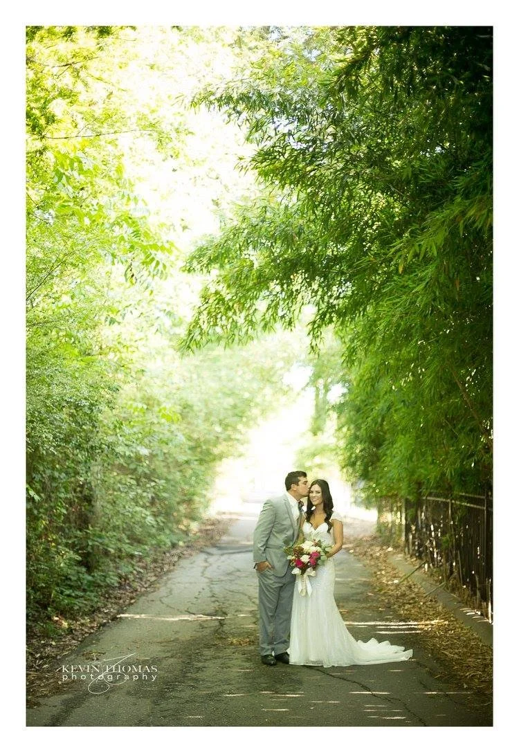 A bride and groom standing on a tree-lined path, with the groom kissing the bride on the forehead. The bride is holding a bouquet of flowers and is wearing a white wedding dress, while the groom is dressed in a gray suit. Bright sunlight is shining t