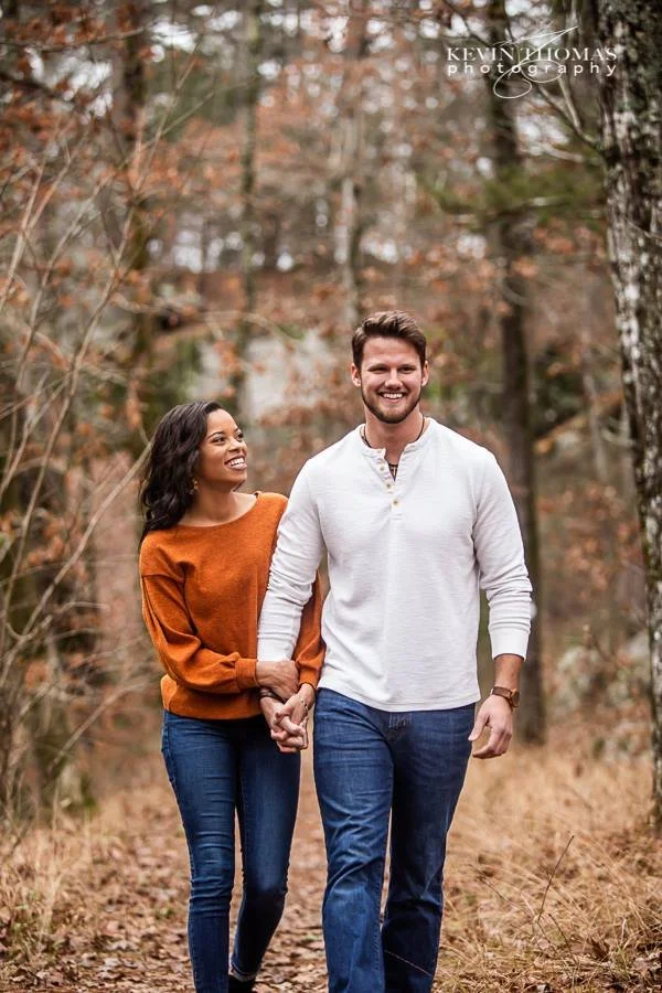 A smiling couple walking hand-in-hand through a autumnal forest with fallen leaves and trees with orange and brown foliage.