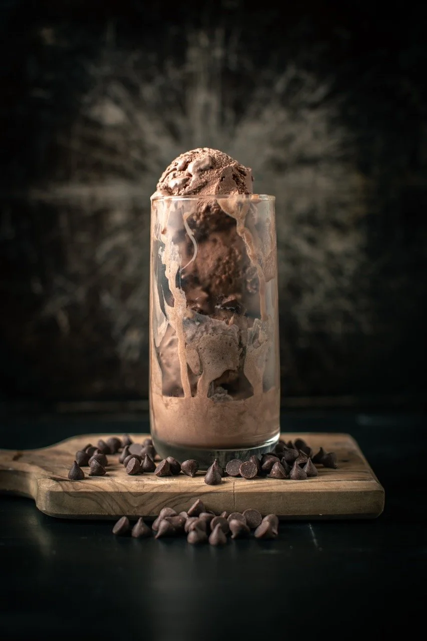 A glass of chocolate ice cream with chocolate chips on a wooden board, with chocolate chips scattered around, against a dark background.