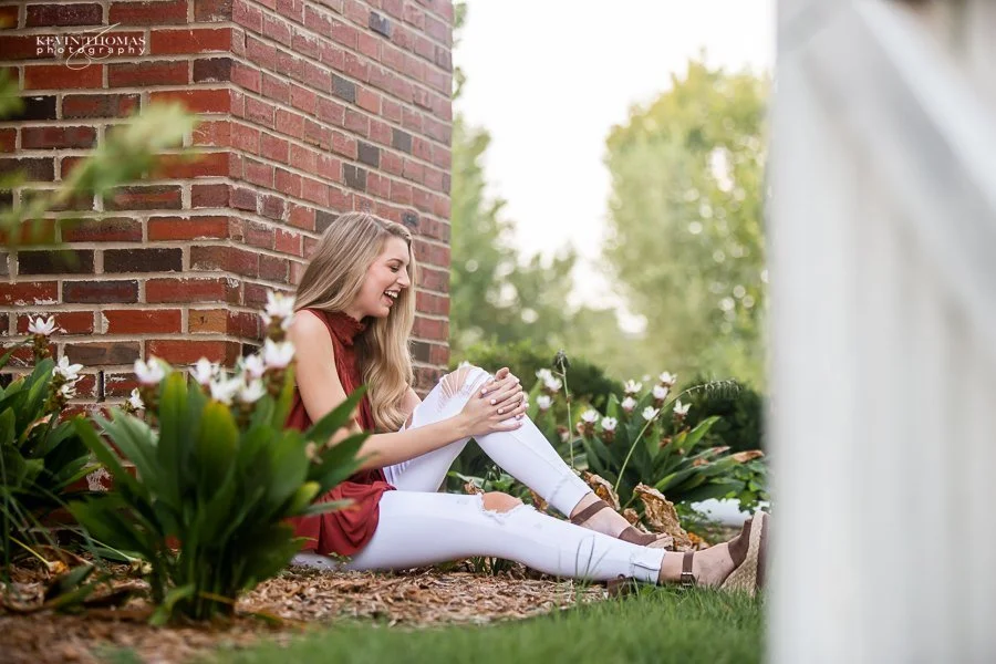A young woman sitting on the ground against a brick building, smiling and laughing, surrounded by green plants and flowers outdoors.