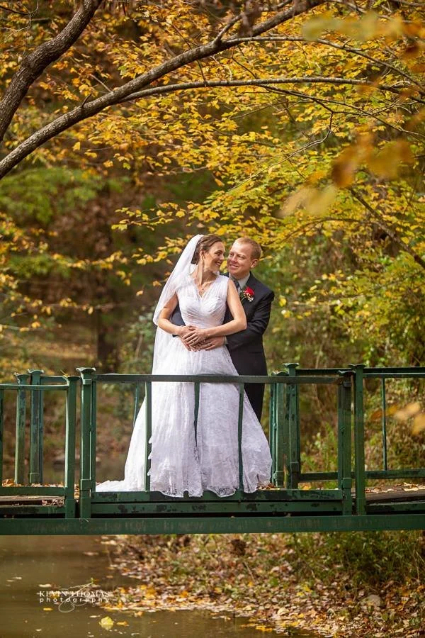 Bride and groom standing on a small green bridge over water with autumn leaves on trees in the background.