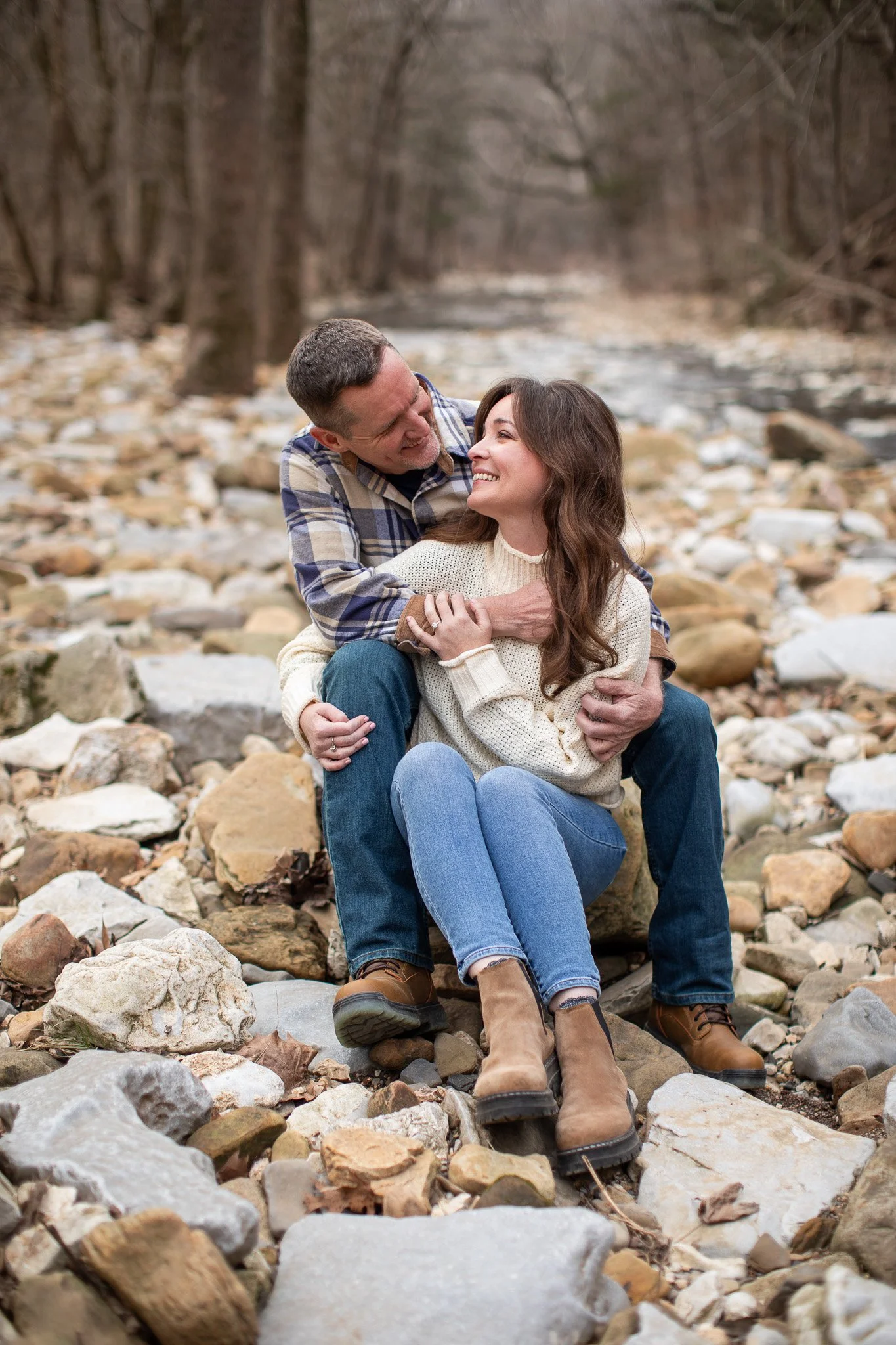 A man and woman sitting on rocks near a river, smiling and enjoying each other's company in a wooded area.