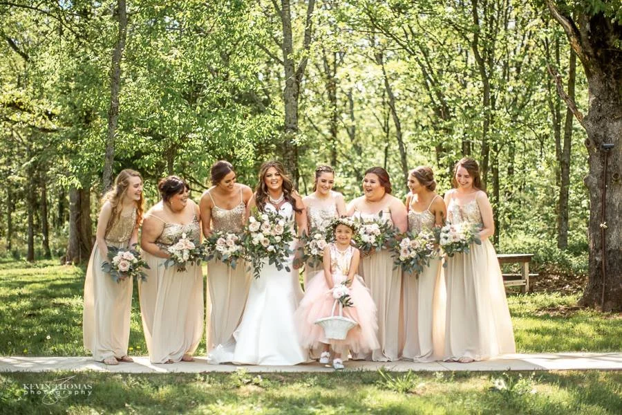 A wedding party outdoors in a wooded area, including the bride, six bridesmaids, and a flower girl with a bicycle, all dressed in formal attire and holding bouquets.