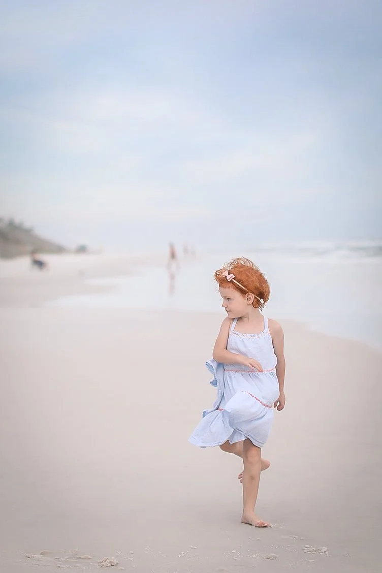 A young girl with red hair, wearing a light blue dress, running barefoot on a sandy beach.