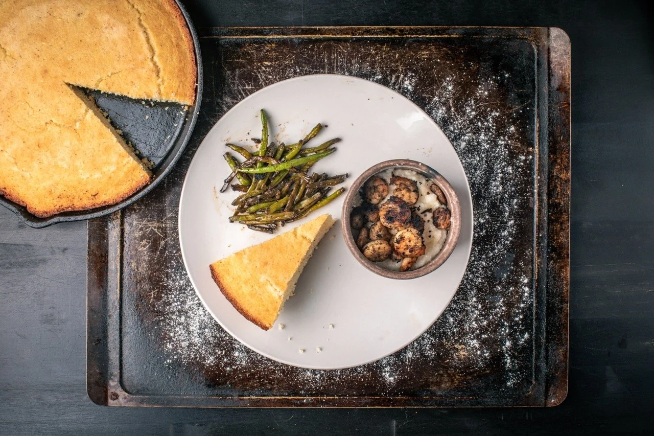 A plate with a slice of cornbread, cooked green beans, and grilled mushrooms, next to a whole cornbread on a black baking tray dusted with flour.