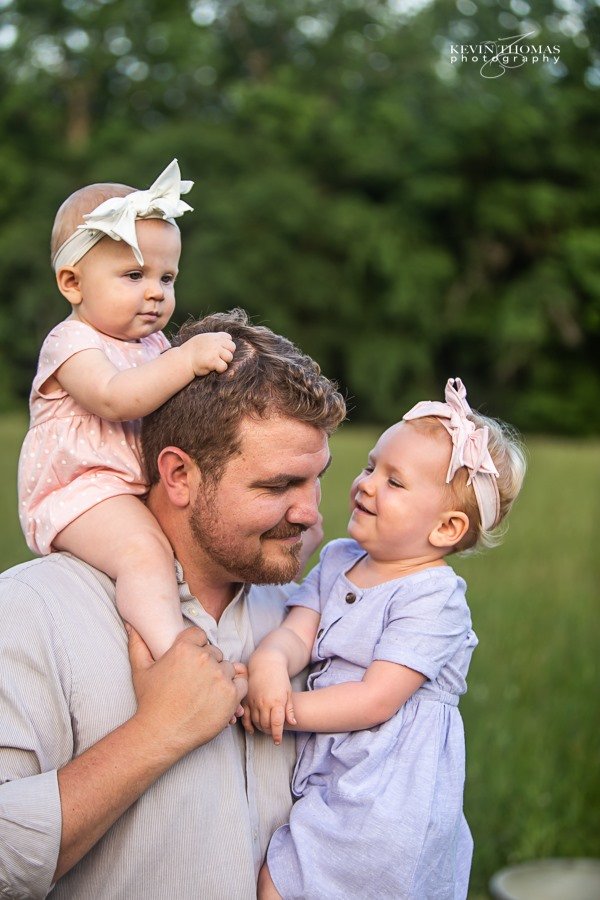 A man with two young girls outdoors in a park, one girl sitting on his shoulders and the other in his arms, both girls wearing headbands and pastel dresses.