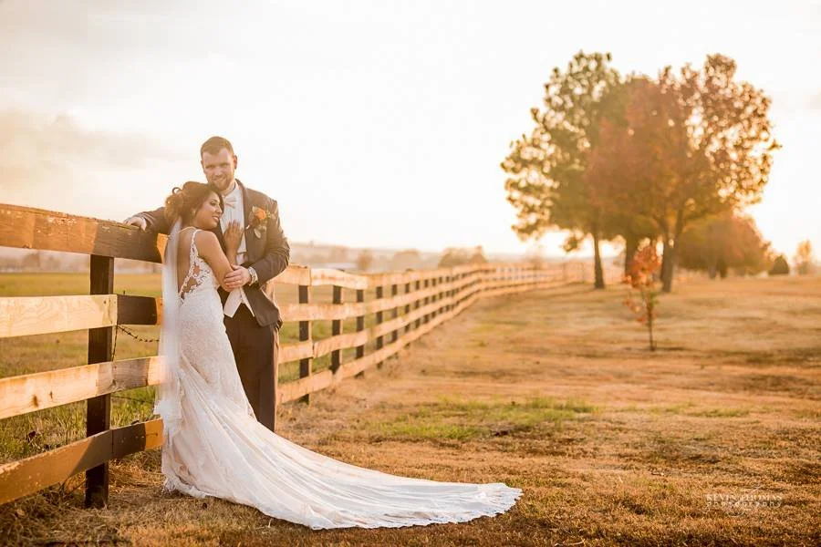 A newlywed couple stands by a wooden fence in a rural field during sunset. The bride is in a white wedding dress, leaning on the groom, who wears a suit with a boutonnière. The scene has warm, golden lighting and a tree in the background.