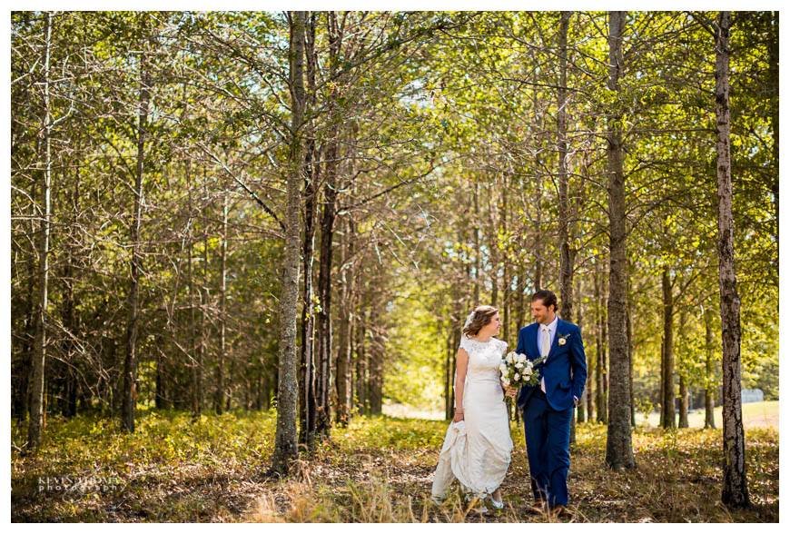 Bride and groom walking hand in hand through a sunlit forest, with trees and green leaves around them. The bride is holding a bouquet of flowers.