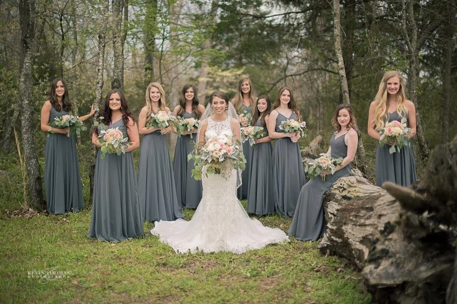 A bride in a white wedding gown with veil and nine bridesmaids in matching blue dresses holding bouquets in a wooded outdoor setting.